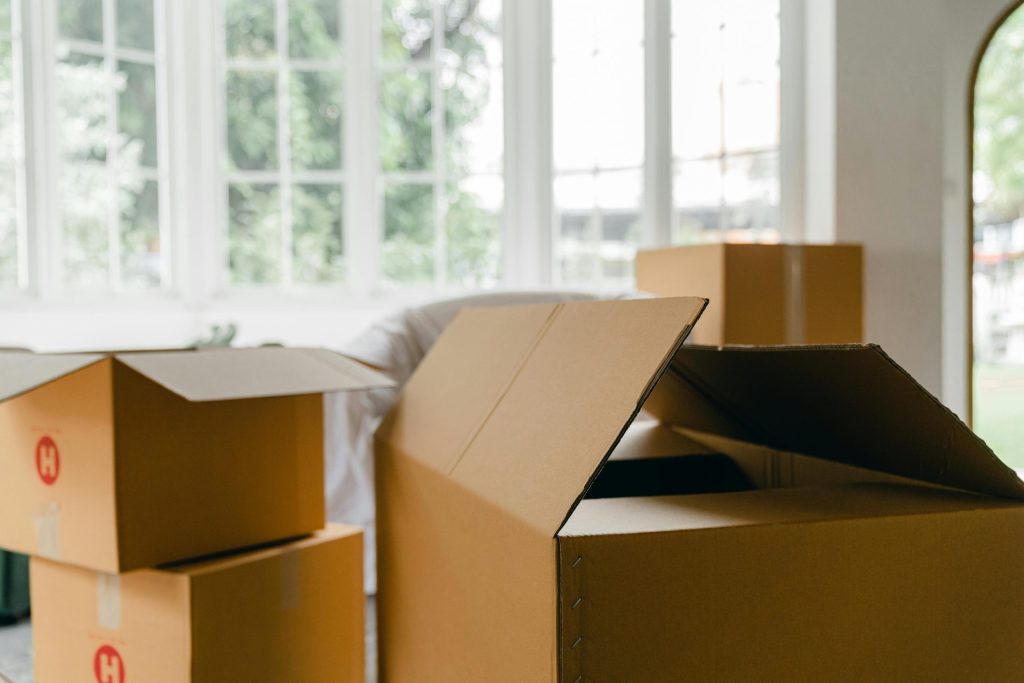 Leere, ungepackte Umzugskartons für persönliche Gegenstände stehen mitten in einem modernen Zimmer neben einem mit einem Laken abgedeckten Sessel vor dem Panoramafenster.